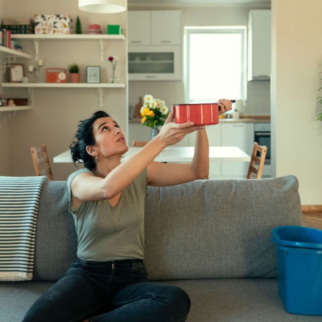 An image of a young woman on her couch whose roof is leaking. She's trying to catch the water.